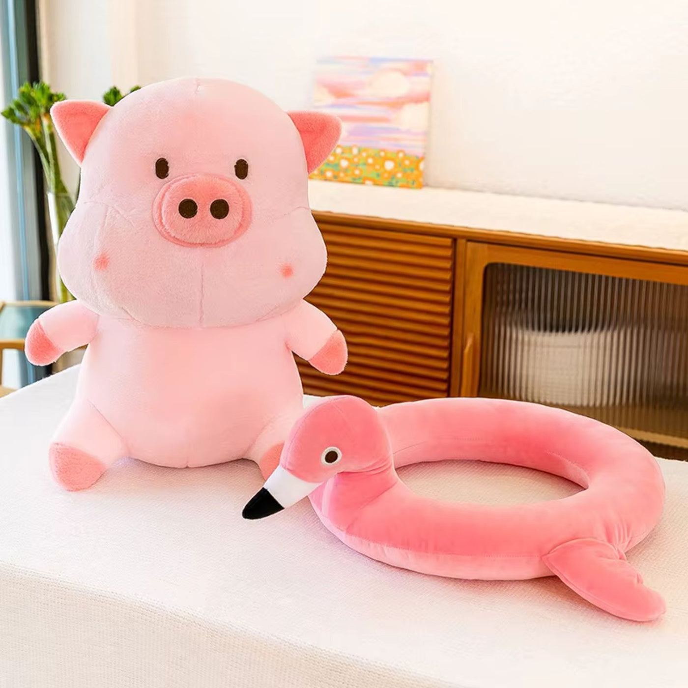 Pink plush pig and flamingo neck pillow on a white surface with a blurred background